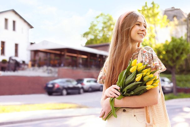 Beautiful teenage girl with bouquet of yellow tulips on city street, space for text - Zdjęcie, obraz