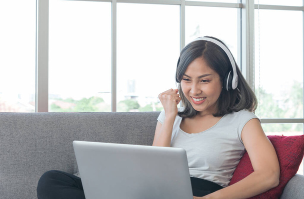 Günlük giysiler içindeki Asyalı genç iş kadını evdeki oturma odasında kulaklık takarak rahatlıyor. Dizüstü bilgisayarla rahat koltukta çalışırken elini kaldırıyorsun. Dünya Sağlık Örgütü. evden çalışma ve başarılı konsept - Fotoğraf, Görsel
