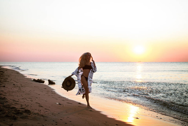 Praca artystyczna sylwetka szczęśliwej dziewczyny w kapeluszu, biegającej wzdłuż piaszczystego brzegu oceanu, w pobliżu wzgórza, rano o świcie. - Zdjęcie, obraz
