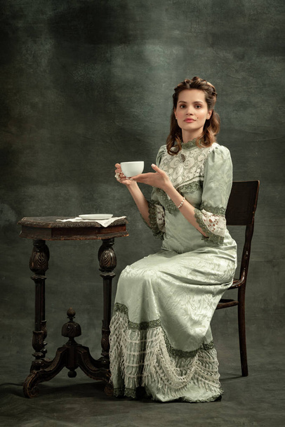 Tea time. Vintage portrait of young beautiful girl in gray dress of medieval fashion style isolated on dark background. Comparison of eras concept, flemish style. Classic art character, old-fashioned. - Foto, Imagem