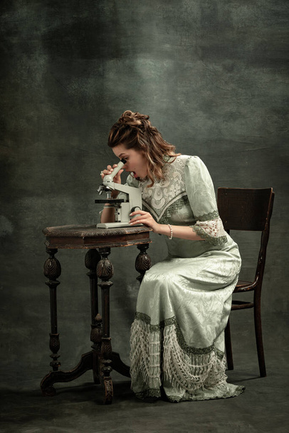 Scientific discovery. One young beautiful girl, actress in image of medieval person using microscope isolated on dark background. Comparison of eras concept, emotions, flemish style. - Photo, Image