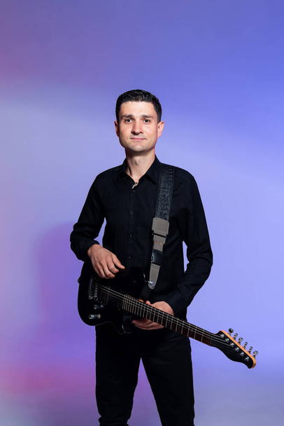 Cheerful guitarist in black clothes with a black electric guitar in his hands. Colorful background - Photo, Image