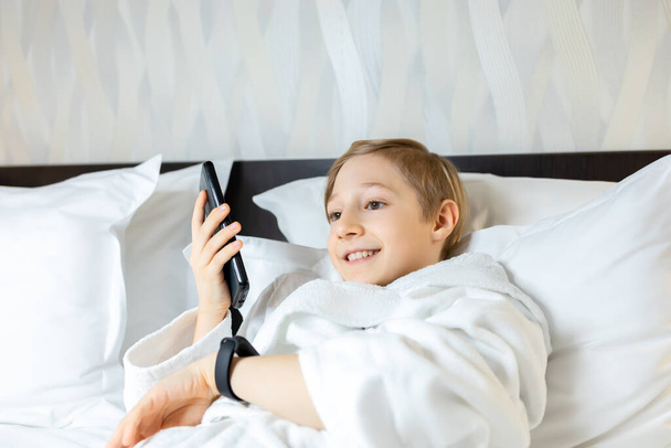 a boy in a hotel room lies on the bed holding a phone in front of him and talking on the speakerphone on the phone - Foto, Bild