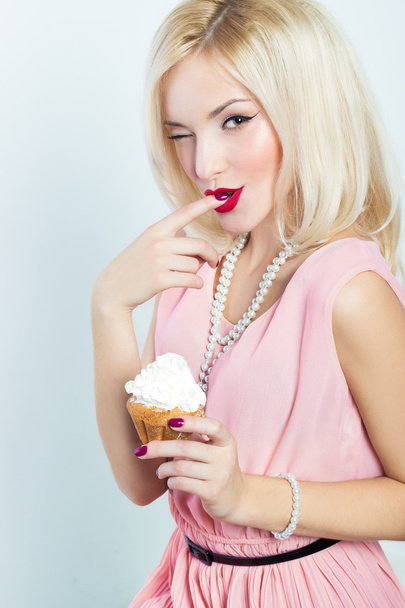 Beautiful sexy elegant blonde woman with bright makeup in pink dress in the Studio on a white background sitting with a cupcake in her hands and licks the sweet cream from his finger winks retro style - Foto, Imagen