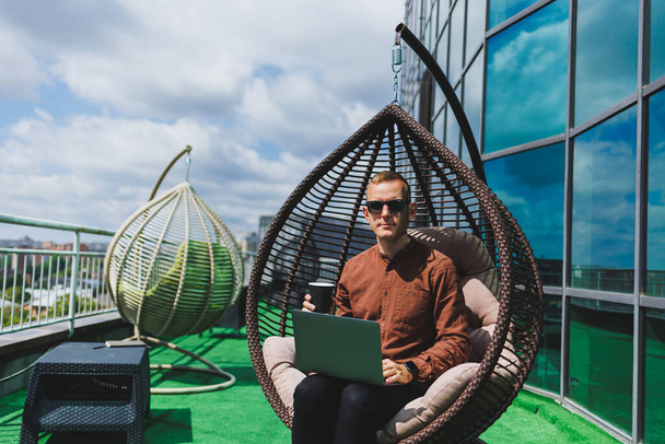 Biznesmen w formalnym ubraniu siedzi na fotelu i patrzy na laptopa podłączonego do sieci bezprzewodowej 4g na tarasie nowoczesnego biura, pracując zdalnie - Zdjęcie, obraz