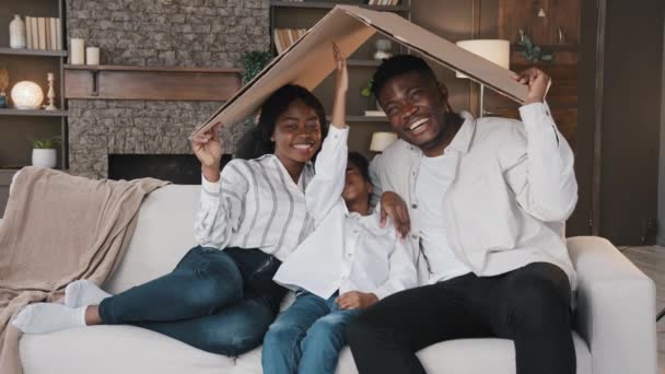 Африканская американская семья родителей с дочерью маленькой девочки усыновленный ребенок сидеть на диване под картонной крышей мечтает о собственном доме переселения жилья страхование недвижимости покупая квартиру ребенка защиты. Высокий - Кадры, видео
