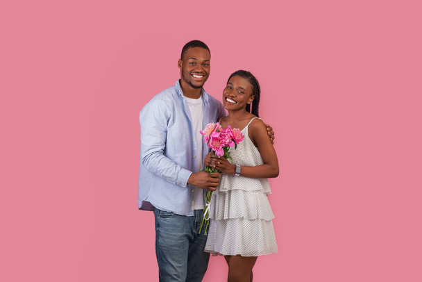 Portrait d'un beau couple noir posant en regardant la caméra, femme tenant un bouquet de fleurs, homme heureux embrassant et saluant dame avec anniversaire ou Saint-Valentin, Journée internationale de la femme, 8 mars - Photo, image
