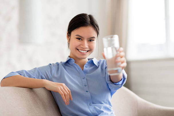Zdravá dobře vypadající mladá Číňanka doporučující pití vody, hezká asijská dáma sedící doma na pohovce a držící sklenici čerstvé vody, usmívající se na kameru, kopírovací prostor, detailní záběr - Fotografie, Obrázek