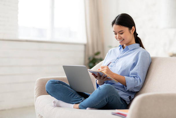 Feliz milenar mulher asiática em roupa casual estudante frequentando webinar de casa, sentado no sofá, usando laptop e tomar notas, tendo curso on-line, vista lateral, espaço cópia. Conceito de E-educação - Foto, Imagem
