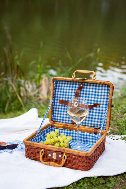 Romantic picnic in the park on the grass against the backdrop of a beautiful lake, delicious food: picnic basket, wine, grapes, figs, cheese, blue checkered tablecloth, two glasses of wine.Outdoor recreation concept - 写真・画像