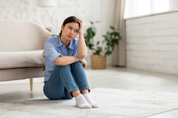 Bored young korean woman in casual outfit sitting on floor by couch at home, touching her head and looking at copy space, suffering from lifestyle crisis, living room interior, panorama - Foto, Imagen