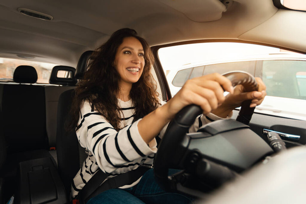 Confident Professional Driver. Portrait of happy young woman sitting in luxury car on drivers seat. Positive black lady riding in the city holding hands on steering wheel, looking at road - Photo, Image
