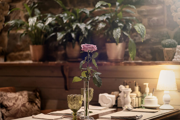 Une rose dans un vase mince transparent se dresse sur une table servie dans un café avec un intérieur de style vintage. Focus sur la rose dans le vase - Photo, image
