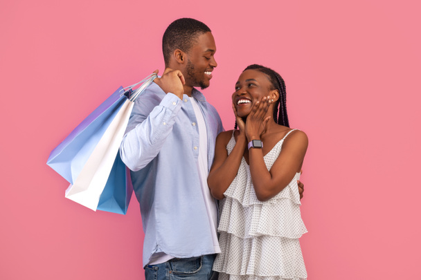 Shopping, Retail, Sales And Discounts. Excited black lady looking at guy, cheerful couple hugging, man holding many bright shopper bags after buying gifts and clothes standing isolated on pink wall - Photo, Image