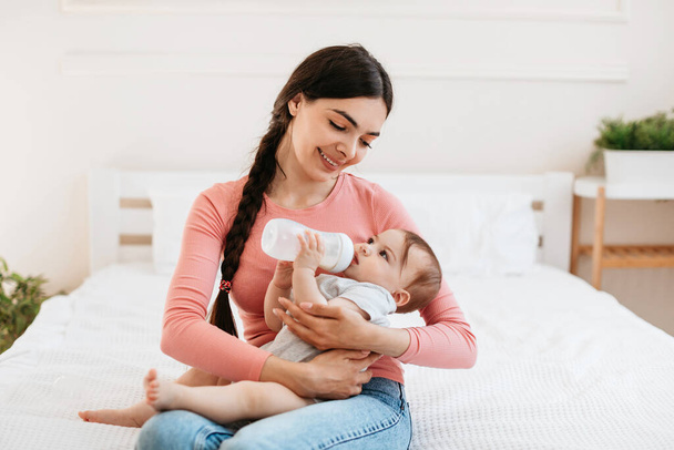 Cute infant baby drinking milk from bottle while lying on mothers hands, thirsty little child enjoying healthy drink. Woman sitting on bed at home - Foto, imagen