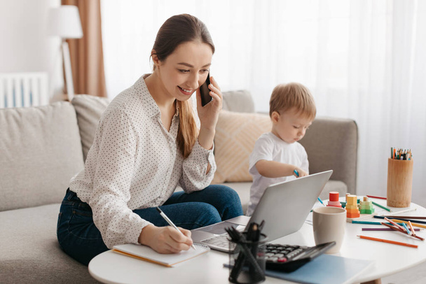 Happy business mother working at home while her little son playing next to her, lady talking on smartphone and taking notes, multitasking and spending time together at home - Foto, afbeelding