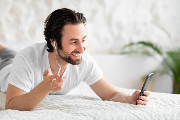 Joyful young guy laying on bed at home, using brand new smartphone and earphones, looking at cell phone screen, smiling and gesturing, having video conference with lover, copy space - Foto, Imagem