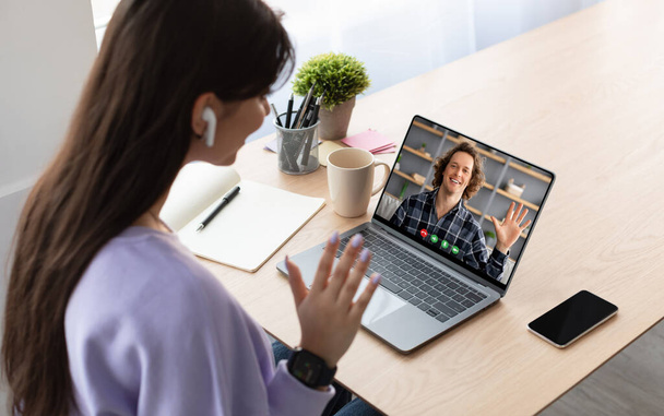 Online Communication, Video Call, Remote Meeting. Over shoulder view of young woman sitting at desk and talking using laptop, waving at webcam. Lady chatting on web with cheerful guy, using earpods - Photo, Image