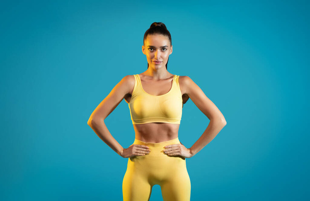 Sarı spor elbiseli bir kadın, kameraya bakıyor, el ele tutuşuyor, mavi arka planda duruyor, stüdyoda çekim yapıyor. Fitness Çalışması ve Motivasyon Konsepti - Fotoğraf, Görsel