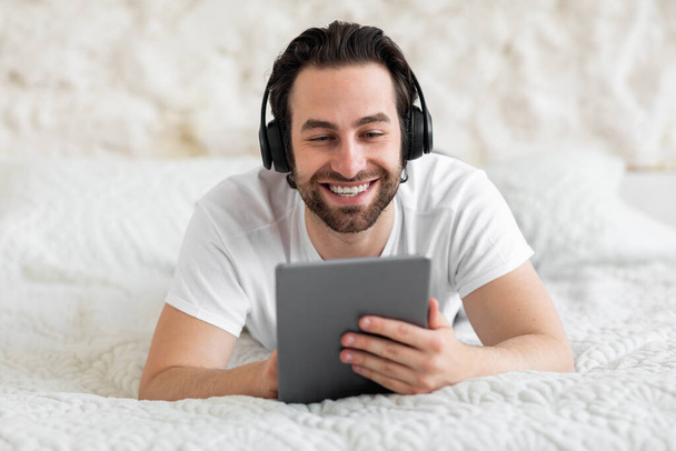 Happy vousatý mladý muž ležící na posteli, pomocí zbrusu nové digitální tablet a bezdrátové stereo sluchátka, sledování videa na internetu při relaxaci v posteli, s videohovory, detailní záběr, kopírovací prostor - Fotografie, Obrázek