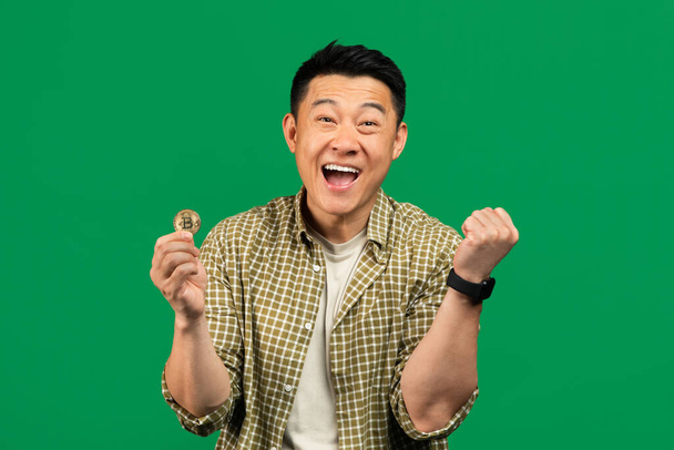 Joyful asian mature man showing yes gesture and holding golden bitcoin in his hand, posing over green background. Advertising of btc crypto coin - Photo, Image