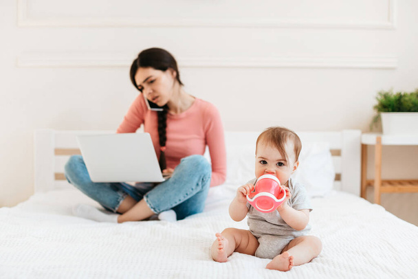 Zajęta matka pracuje na laptopie i rozmawia przez telefon, podczas gdy jej dziecko pije wodę z butelki, siedzi w domu w łóżku. Tysiącletnie kobiety pracujące na urlopie macierzyńskim - Zdjęcie, obraz
