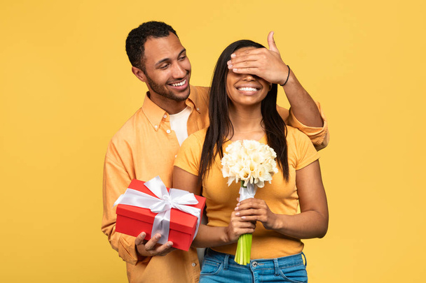 Όμορφος νεαρός μαύρος δίνει λουλούδια και δώρα στην κοπέλα του, καλύπτει τα μάτια της, κάνοντας έκπληξη γενεθλίων στο κίτρινο φόντο του στούντιο. Ρομαντικό εορτασμό, έννοια των διακοπών - Φωτογραφία, εικόνα