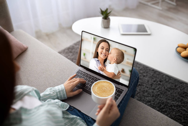 現代の遠隔通信の概念。若い女性の肩のビューの上に作るオンラインビデオ通話で女性と赤ちゃんの子供,ソファにノートパソコンとコーヒーを飲みながら座って,コピースペース - 写真・画像