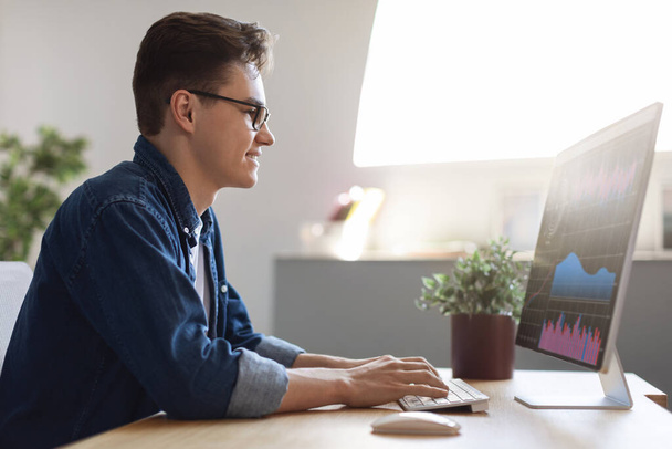 Profilbild eines jungen Geschäftsmannes, der am Computer am Arbeitsplatz im Büro arbeitet, männlicher Unternehmer, der am Schreibtisch sitzt, Monitor mit Online-Handelsstatistiken betrachtet und auf Tastatur tippt, freier Platz - Foto, Bild