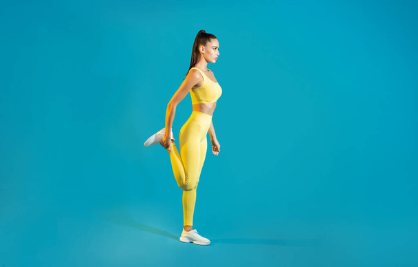 Fitness Lady Stretching Leg And Hamstring Muscles Warming Up Before Workout Standing On Blue Studio Background, Wearing Yellow Fitwear. Side View, Full Length Shot - Photo, image