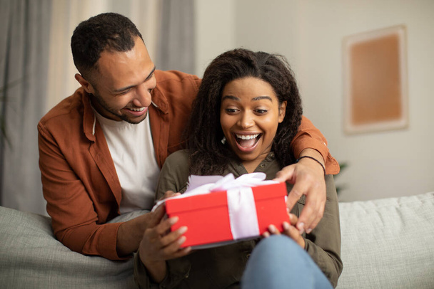 Happy Black Husband Giving Gift Box To Joyful Wife Congratulating Her And Celebrating Family Holiday Sitting On Sofa At Home. Celebration And Present Delivery Concept - Photo, Image