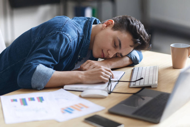 Overwork Concept. Exhausted male employee sleeping at desk in office, tired millennial man napping at workplace, leaning at table with head placed on hands, lying on opened notepad, closeup shot - Foto, afbeelding