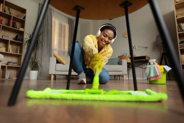 Afro-amerikan bayan paspas yapıyor kulaklıkla müzik dinliyor evde oturma odasını temizliyor. Ev Temizliği ve Hizmetçi Hizmetleri Konsepti. Seçici Odaklanma - Fotoğraf, Görsel