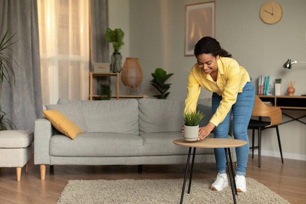Afroamerikanerin stellt Pflanzenkübel auf den Tisch, putzt und dekoriert Haus im modernen Wohnzimmer zu Hause. Innenarchitektur, Dekoration und häusliche Chores Konzept. Volle Länge - Foto, Bild