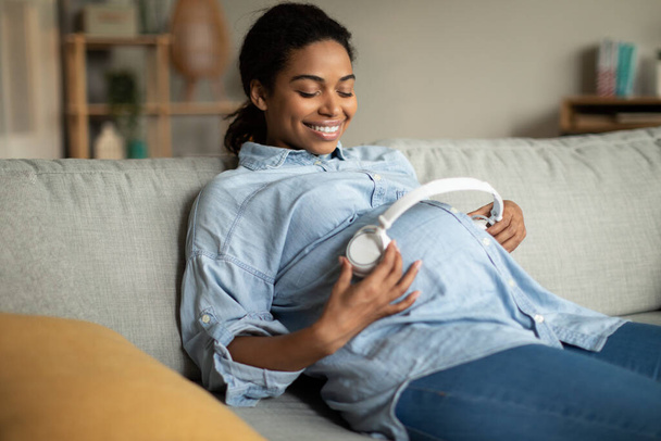Happy Pregnant Black Woman Listening To Music Online Holding Wireless Headphones Near Belly Sitting On Couch At Home. Pregnancy Leisure, Childbirth Concept - Foto, imagen
