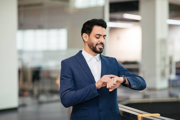 Happy confident young islamic ceo manager with beard in suit looks at smart watch in office interior, wait meeting. Nodern online gevice for successful business, boss career, lifestyle and leadership - Photo, Image