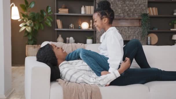 African mother babysitter lying on sofa playing with biracial child playful daughter tickle girl on couch laughing having fun tickling smiling together. Mum and kid enjoying holiday play game relax - Footage, Video