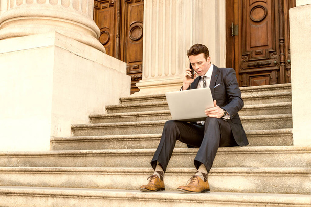 American Businessman traveling, working in New York, dressing in black suit, leather shoes, sitting on stairs outside office, working on laptop computer, calling on phone.  - Photo, Image