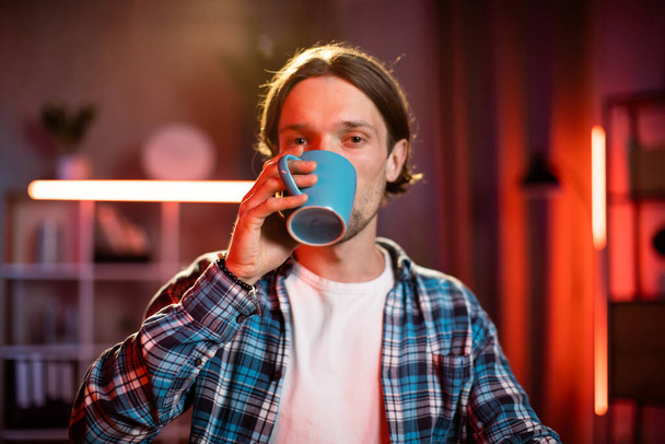 Close up of hand some young blond man with a hot drink in a blue mug wearing checkered shirt. Professional male worker in casual wear, smiling while looking at camera - Фото, зображення