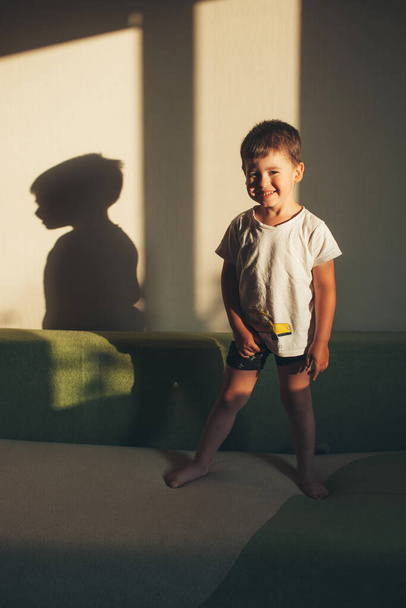 Porträt eines Jungen, der auf dem Sofa sitzt und eine Spielpause einlegt, um ein Foto zu machen. Junge steht auf Sofa. Lächelndes glückliches Kind. - Foto, Bild