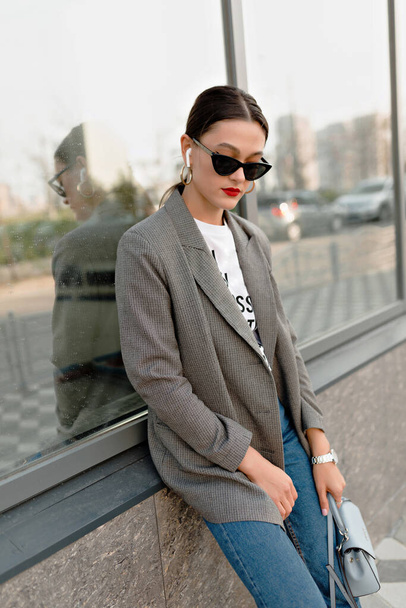 Fashionable woman in sunglasses wearing grey jacket and jeans walking in city and listening music. Pretty European woman spend time outside  - Fotó, kép