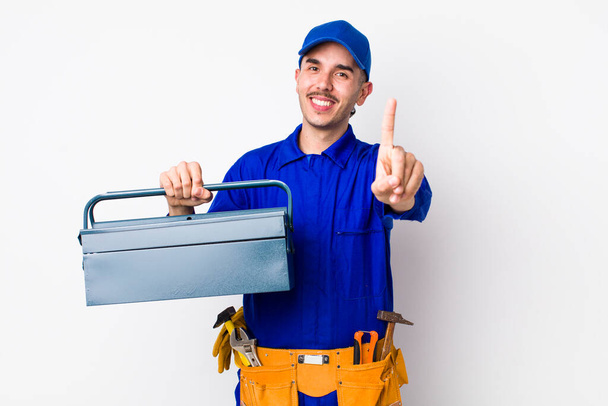 young hispanic plumber smiling proudly and confidently making number one. toolbox concept - Foto, afbeelding