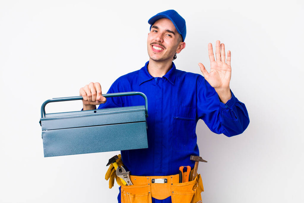 young hispanic plumber smiling happily, waving hand, welcoming and greeting you. toolbox concept - Foto, Imagem