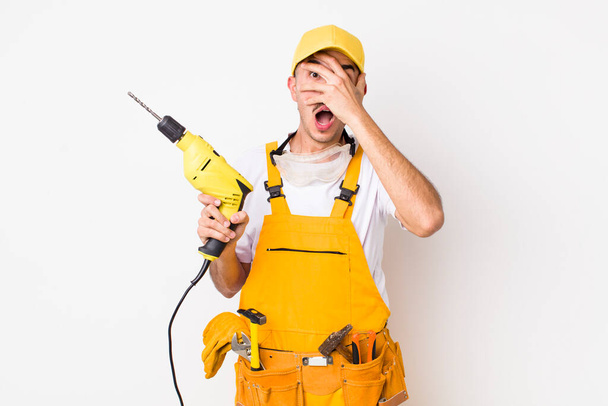 hispanic handyman looking shocked, scared or terrified, covering face with hand. drill concept - Foto, Bild