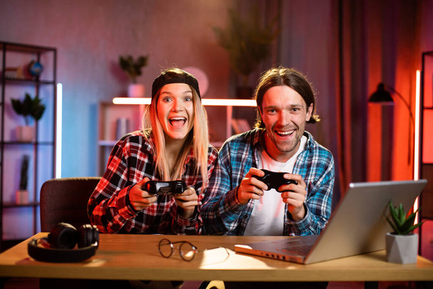 Young multiracial couple sitting at table with wireless joysticks in hands and playing video games on laptop. Caucasian man and blond woman in black cap spending evening time at home with fun. - 写真・画像