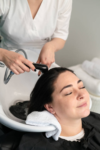 Kozmetik uzmanı ameliyatlar için kızın kafasını şampuanla yıkar. Siyah saçlı. artı boyut. - Fotoğraf, Görsel
