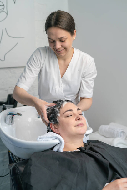 косметолог промывает девушке голову шампунем для дальнейших процедур. черные волосы. плюс размер. - Фото, изображение