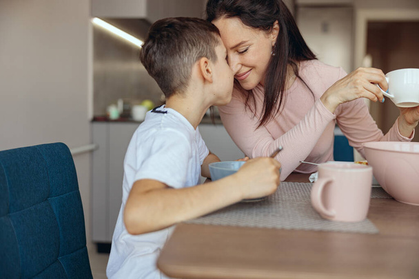 Boldog anya és kis tini fiú összeölelkeznek a konyhában és esznek. Jó reggelt! Anya szeret. - Fotó, kép