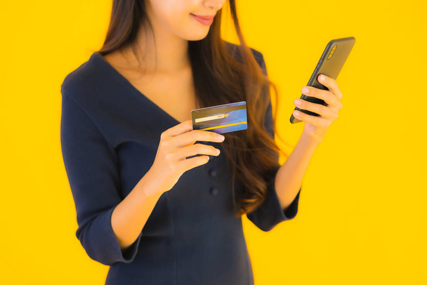 Retrato hermosa mujer asiática joven con teléfono móvil inteligente o teléfono celular y tarjeta de crédito amarillo aislado fondo
 - Foto, Imagen