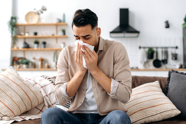 Unhappy sad indian or arabian guy suffering from fever and flu, sitting on sofa in living room, blowing nose and sneezing in napkin, need treatment of illness, and doctors consultation - Фото, зображення